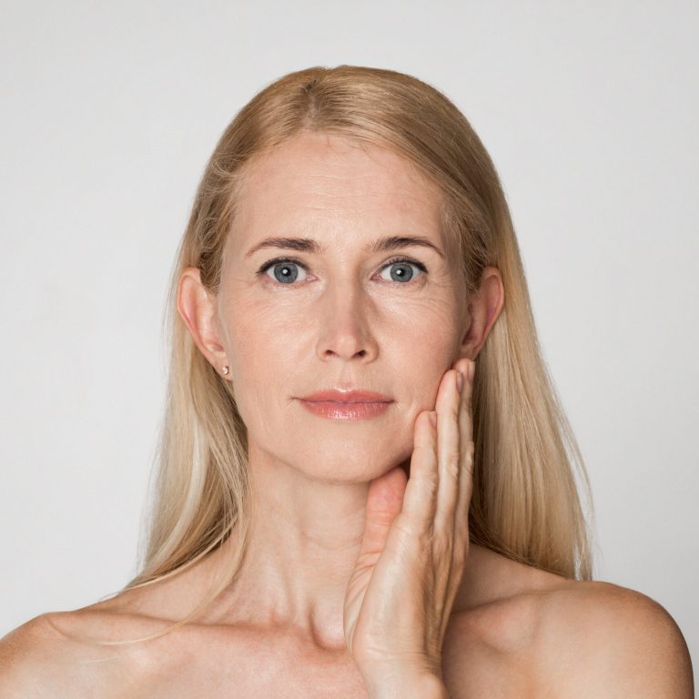 A middle aged woman with blonde hair. She is looking directly at the camera with a neutral expression. She is touching her cheek with her right hand. The background is a plain white wall.
